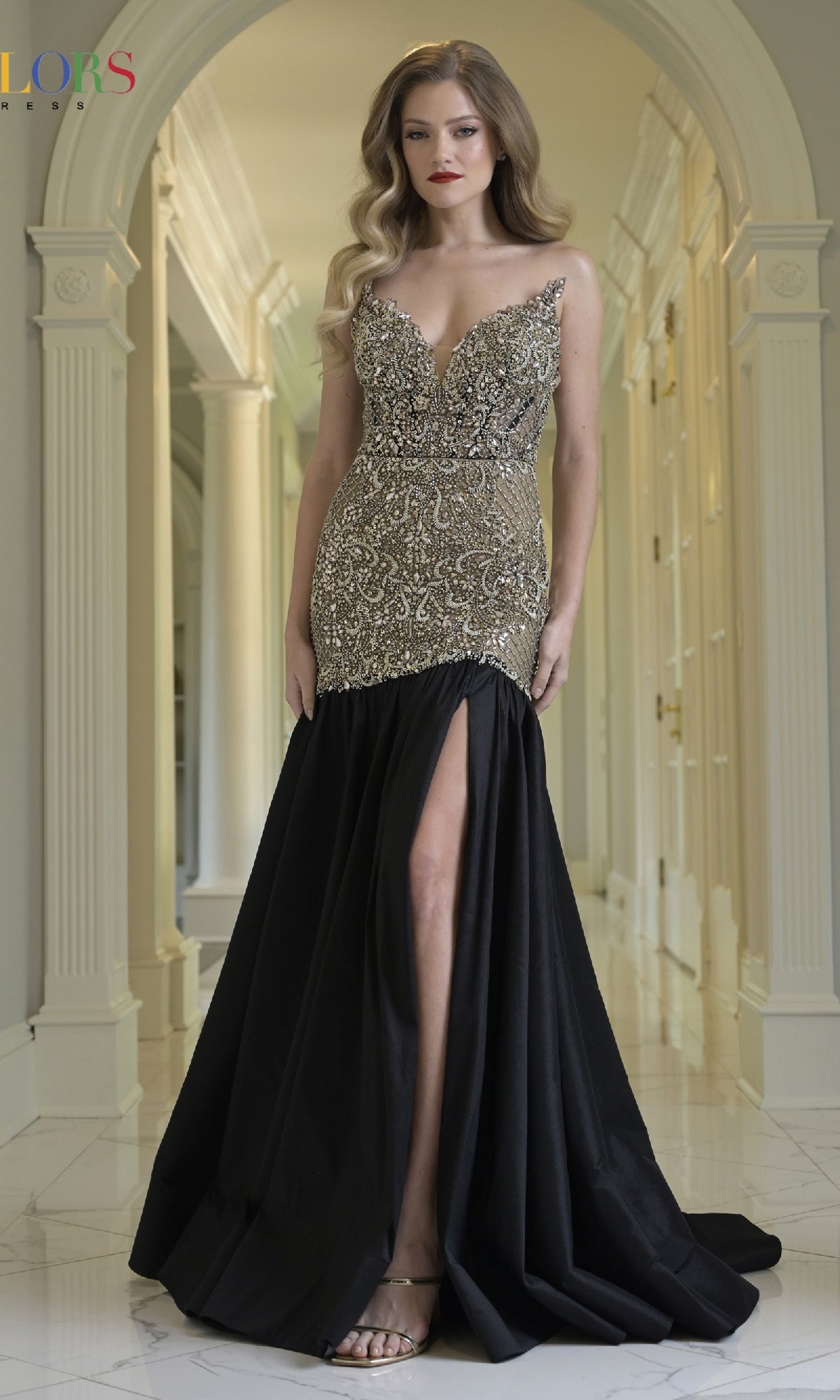 A woman stands in a hallway wearing the Long Prom Dress: Colors Dress 3898, featuring a sleeveless beaded bodice with a plunging neckline and sequin details, plus a high slit in the satin black A-line skirt.